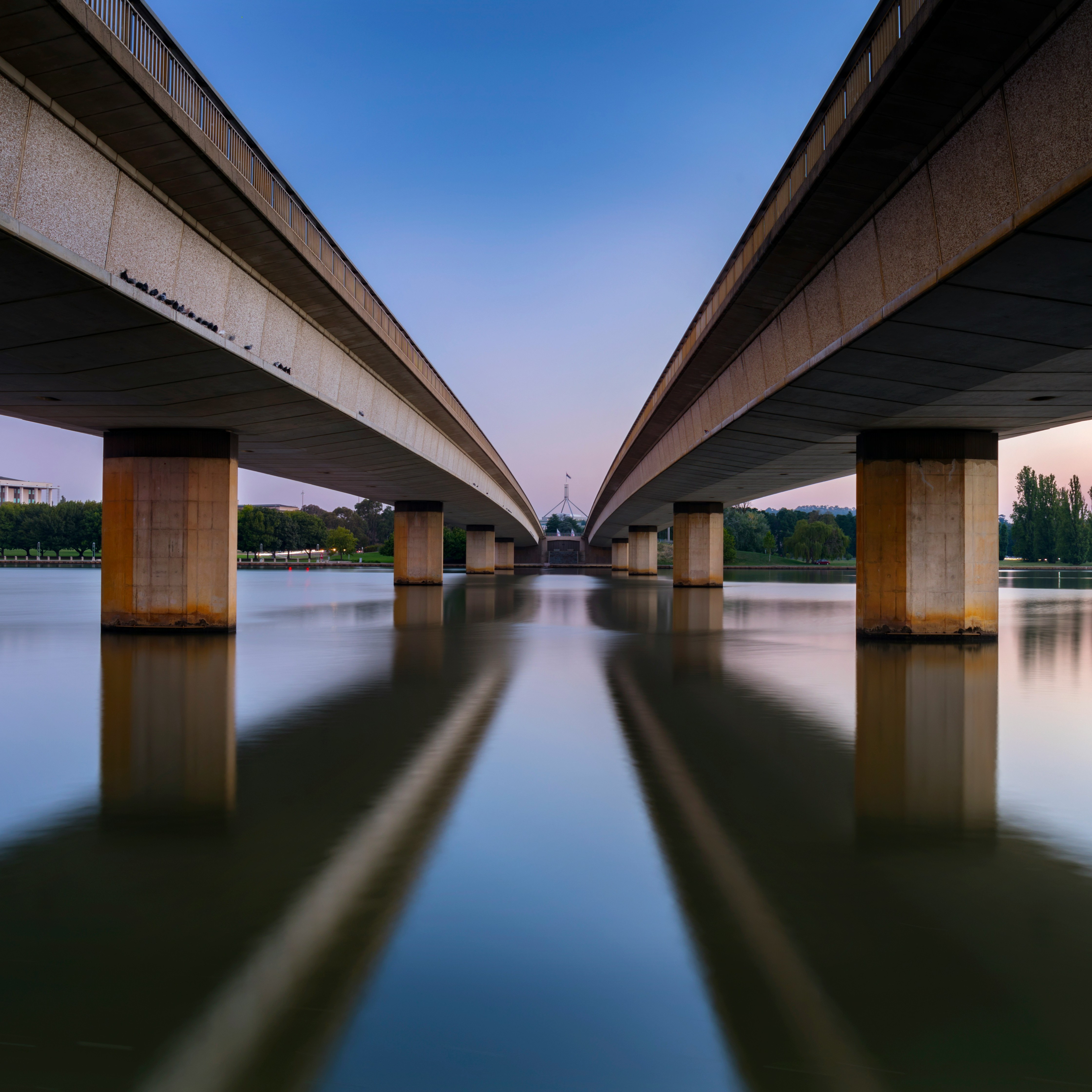 Canberra View Symmetrical view beneath two parallel bridges over calm water at dusk, with Canberra’s Parliament House visible in the distance between the bridge spans.
